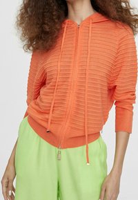 Orange ribbed zip-up hoodie with a hood and drawstrings; features a textured design. Paired with loose-fitting bright green pants.