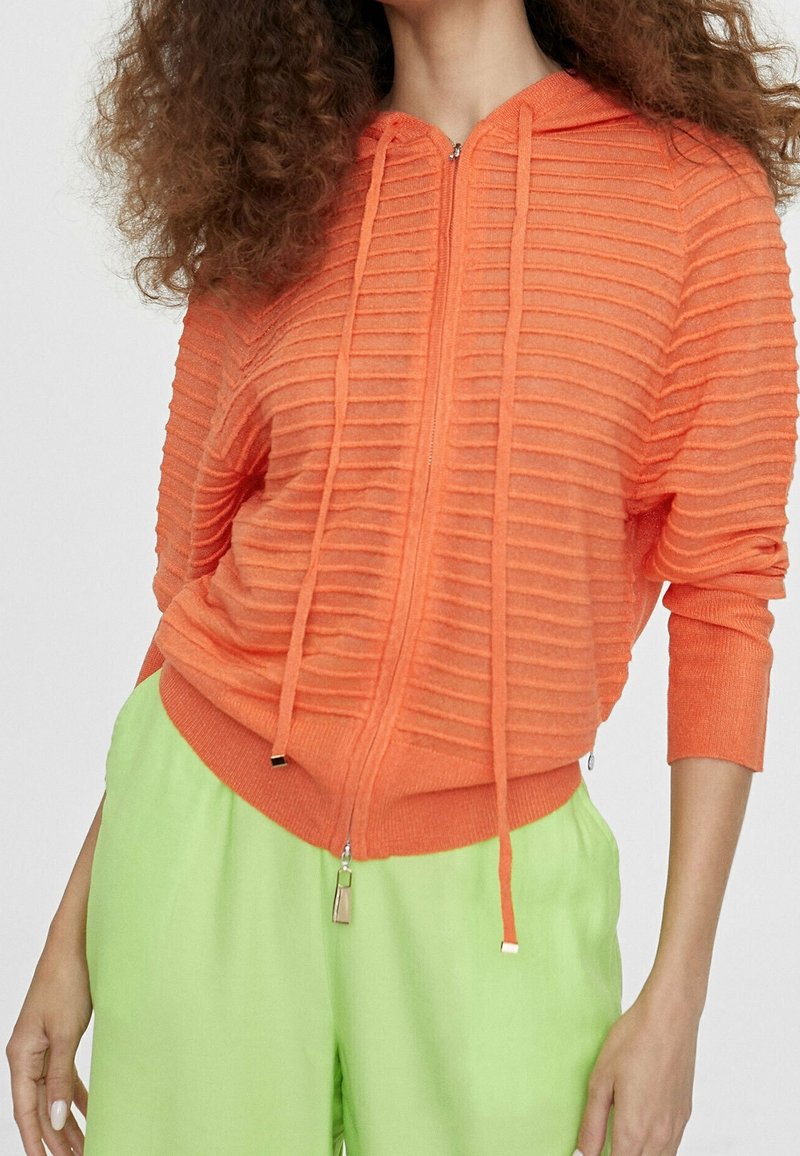 Orange ribbed zip-up hoodie with a hood and drawstrings; features a textured design. Paired with loose-fitting bright green pants.