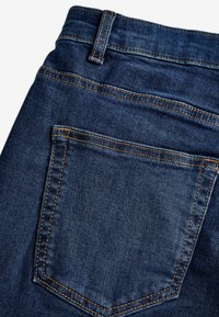 Vaqueros de mezclilla azul oscuro con un bolsillo trasero prominente con costuras naranjas, tejido ligeramente texturizado y un corte clásico a la altura de la cintura.