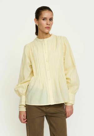Lysegul skjorte med knapper, plisseret front, rund halsudskæring og puffede ærmer. Stoffet er tekstureret med fine syningsdetaljer.