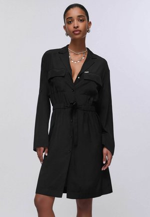 Kobieta nosząca czarną sukienkę koszulową z paskiem, długimi rękawami, z guzikami i warstwowymi srebrnymi naszyjnikami na jednolitym jasnym tle.