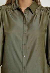 Olive green blouse made of smooth, lightweight fabric, featuring a button-down front and slightly puffed sleeves with gathered details.