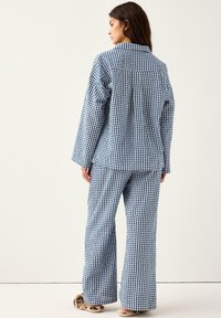 Kobieta stojąca bokiem, ubrana w niebieskie i białe, luźne piżamy w kratę oraz futrzane kapcie, na tle jasnego jednolitego tła.