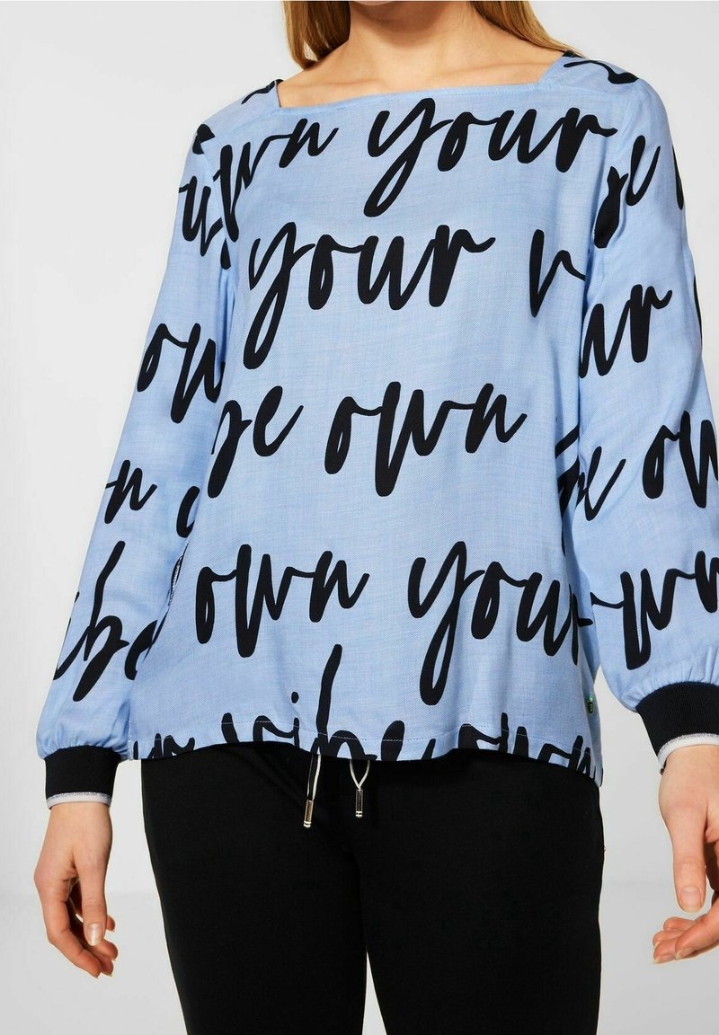 Haut à manches longues bleu clair avec texte manuscrit noir, encolure carrée, manches élastiques et ourlet avec cordon de serrage ajustable.