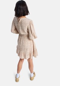 Robe beige avec un motif floral, manches ballon, taille cintrée et ourlet volé. Associée à des chaussures beiges et des chaussettes blanches. Vue de dos.