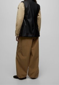 Gilet en cuir noir par-dessus un haut beige, associé à un pantalon large couleur sable. Détails de boutons et de poches visibles sur le gilet et le pantalon.