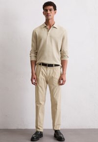 Beige Langarm-Polo mit Kragen, kombiniert mit hellbeigen, plissierten Hosen und schwarzen Lederschuhen. Schlichtes Design und leichte Textur.