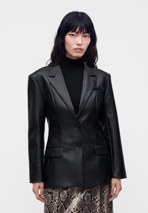 Pessoa com cabelo longo e preto a usar um blazer de pele preto por cima de um top preto e uma saia padrão com design de estampa de cobra.