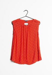 Rode mouwloze blouse van zachte stof met witte stippen, met een knoopsluiting aan de voorkant en geplooide halslijn. Hangt aan een zwarte hanger.