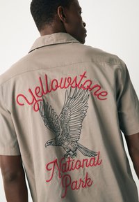 Chemise manches courtes beige avec un dessin au dos comportant le texte brodé rouge « Yellowstone » et une illustration détaillée d’un aigle noir.