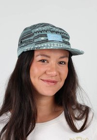 Casquette bleu et gris à motifs avec une visière plate, arborant un patch logo central. Fabriquée en tissu avec des motifs géométriques superposés et des évents latéraux.