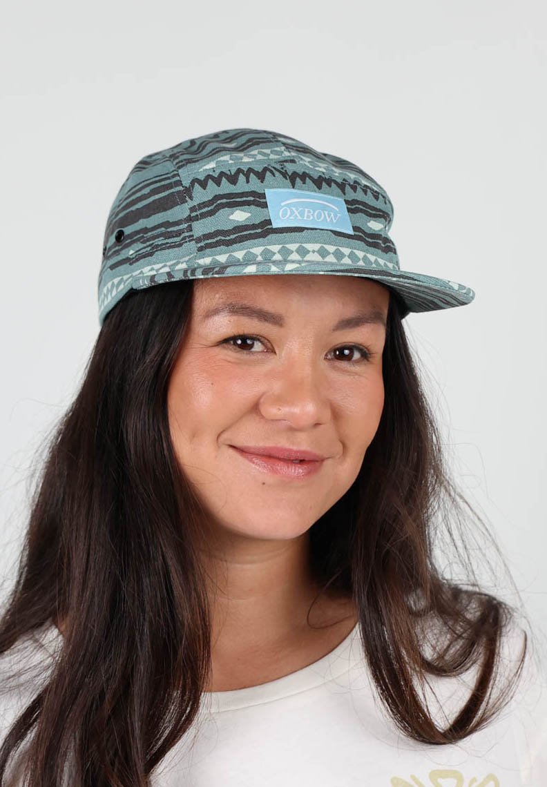 Casquette bleu et gris à motifs avec une visière plate, arborant un patch logo central. Fabriquée en tissu avec des motifs géométriques superposés et des évents latéraux.