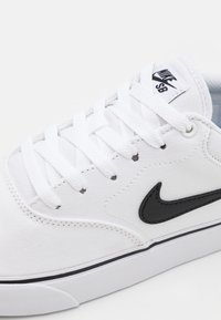 Vit canvas sneakers med svart läder Nike-logotyp, vita snören, perforerad tå och en svart accentlinje längs sulan.