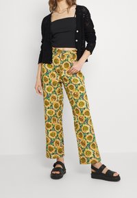 Pantalones acampanados de tela amarilla con un estampado de girasoles. Combinados con un top corto negro y sandalias de plataforma negras. Cárdigan de punto negro.