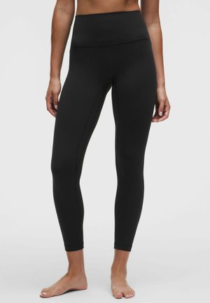 Donna che indossa leggings neri a vita alta e lunghezza totale, in piedi a piedi nudi su uno sfondo chiaro e uniforme.