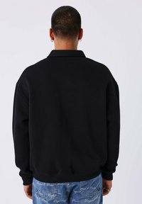 Zwarte, oversized sweatshirt met een omgeslagen kraag, voorzien van een geribde onderrand en manchetten. Zachte textuur, gezien van de achterkant. Gecombineerd met lichtblauwe jeans.