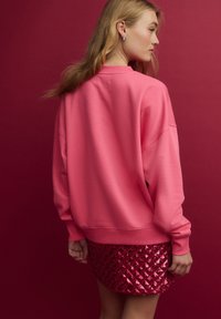 Rožinė per didelė džemperis minkštu audiniu, derinama su raudona pailga sijonu, padengtu deimanto raštu iš blizgučių. Fonas yra vientisas magentinis.