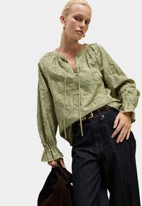 Lichtgroene geborduurde blouse met lange mouwen, ruches aan de manchetten en een V-hals, gecombineerd met donker denim broek en een bruine tas.