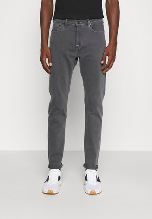 Uomo che indossa jeans aderenti grigio scuro con orli risvoltati, camicia nera e scarpe da ginnastica bianche con dettagli scuri, in piedi davanti a uno sfondo semplice.