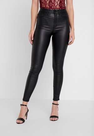 Woman wearing high-waist black faux leather skinny pants, burgundy lace top, and black ankle-strap high-heeled sandals, standing against white background.