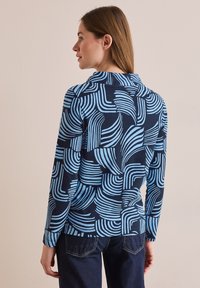 Námořnický top s dlouhým rukávem, který má modrý abstraktní vzor křivolakých linií. Měkká tkanina s límečkem a mírně přiléhavý střih.