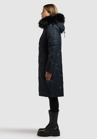 Marineblaue Steppjacke mit Kapuze aus Kunstfell, langer Silhouette und Seitentaschen. Kombination mit schwarzen Kampfstiefeln. Glatte, glänzende Oberfläche.