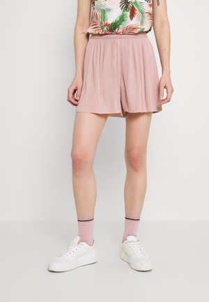 Personne portant un short plissé rose clair, des chaussettes roses à rayures rouges et noires, des baskets blanches et un haut sans manches à motifs floraux, debout contre un mur blanc.