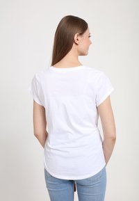 Hvid bomulds T-shirt med korte opsmøgede ærmer, rund halsudskæring og en let buet kant. Modellen har blå jeans på.