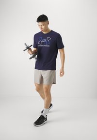 Under Armour sötétkék póló logóval, bézs rövidnadrág, fekete súlyzókat tart, fekete sportcipőt és fehér zoknit visel, semleges háttér előtt.