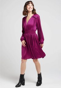 Robe violette avec des manches longues, décolleté en V profond, taille smockée et jupe fluide. Associée à des bottines noires à la cheville. Tissu léger et lisse.