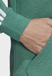 Sudadera verde con un tejido texturizado, acentos de franjas blancas en las mangas y un bolsillo canguro. Primer plano de la mano en el bolsillo.