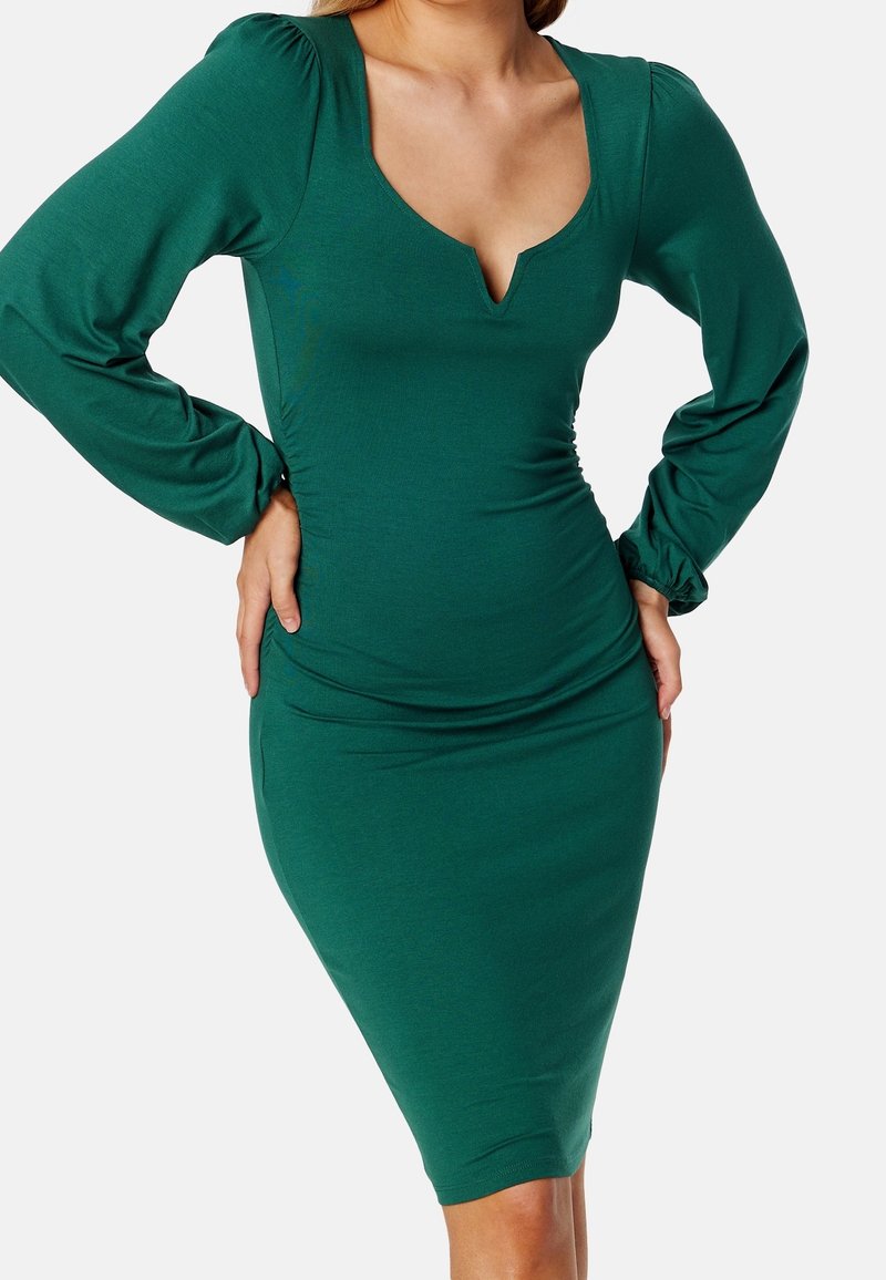 Robe verte moulante avec des manches longues, un décolleté en V profond et des détails froncés à la taille. Fabriquée en tissu lisse et extensible.