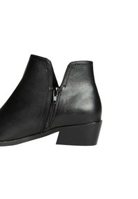 Botín negro de tobillo hecho de cuero suave con cremallera lateral, que presenta un tacón bajo y un diseño en forma de V alrededor del tobillo.