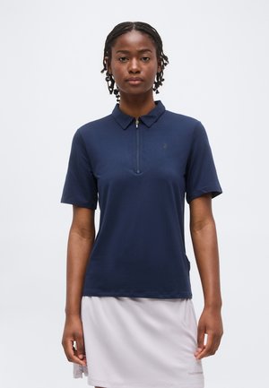 Kvinde med kort fletning iført en mørkeblå kortærmet lynlås-skjorte og et lysegråt skørt, stående mod en ensfarvet baggrund.