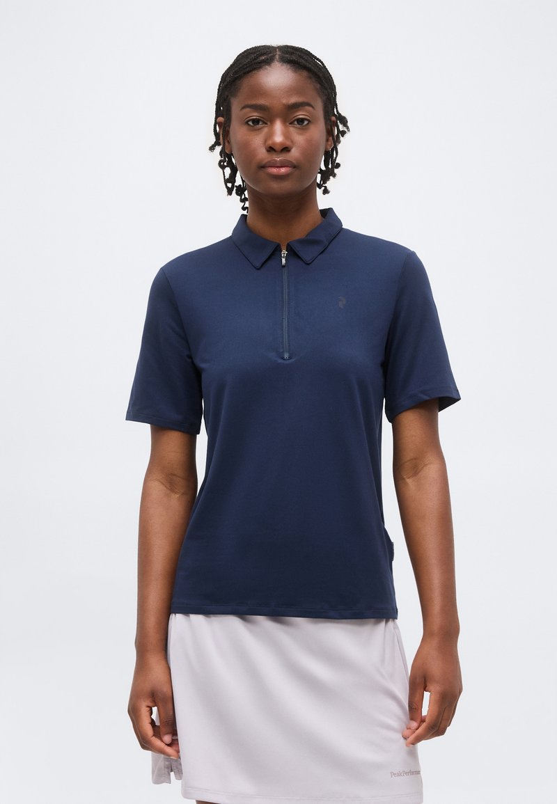 Γυναίκα με κοντά πλεξουδάκια, που φοράει μπλε μπλούζα με φερμουάρ και κοντά μανίκια και ανοιχτό γκρι φούστα, στέκεται μπροστά σε μονόχρωμο φόντο.