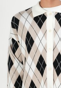 Argyle-patterned cardigan made of soft knit fabric. Features a white collar, button closure, and a color palette of beige, black, and white.