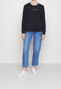 Felpa nera con "HILFIGER" ricamato in testo multicolore; abbinata a jeans dritti blu e sneakers bianche.