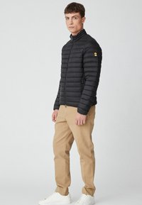 Giacca a vento nera con cuciture orizzontali, colletto alto e chiusura con zip. Abbinata a pantaloni beige e sneakers bianche. Sfondo neutro.