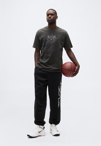 Άνδρας που στέκεται φορώντας μπλούζα NBA All-Star και μαύρη φόρμα, κρατώντας μια μπάλα μπάσκετ στο ένα χέρι, μπροστά σε απλό φόντο.