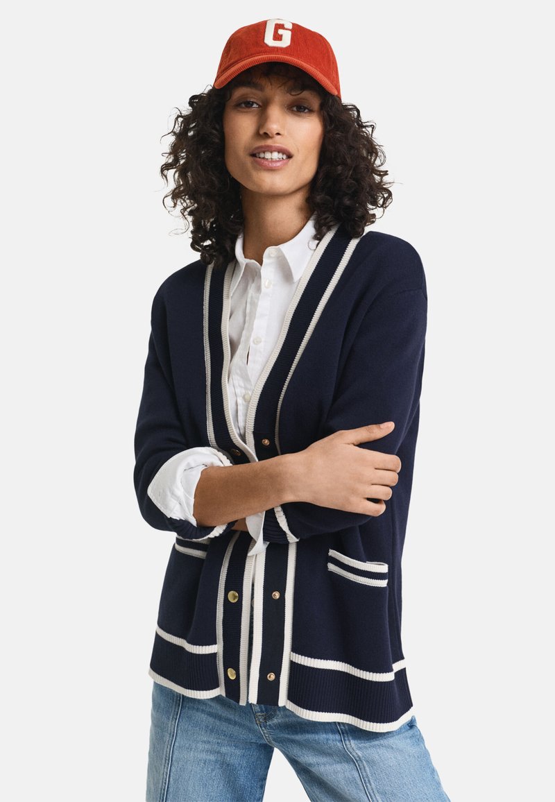 Cárdigan azul marino con rayas blancas, con botones dorados y bolsillos. Lleva puesto sobre una camisa blanca con cuello, combinado con vaqueros claros.