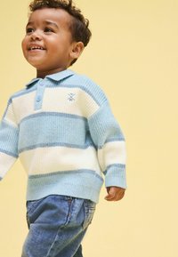 Vaaleansininen ja valkoinen raidallinen villapaita, resoripintainen, kauluksellinen kahdella napilla, yhdistetty sinisiin farkkuihin. Yksinkertainen tausta.