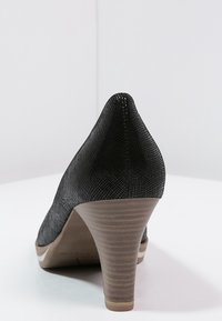 Svart texturerad högklackad sko med spetsig tå, som har en tan-färgad sula och en räfflad klack. Rent design med minimala detaljer.