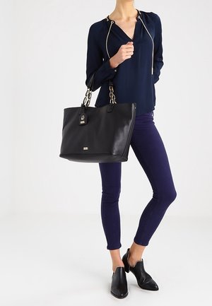 Zwarte leren tote bag met een pebbled textuur, silver hardware, bovenhandvatten en merketiket. Model draagt een marineblauwe blouse en donkere skinny jeans.