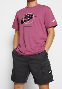 Lila bomull t-shirt med en stor svart Nike-logotyp och text, tillsammans med svarta cargo shorts med en sidoficka med dragkedja och logotypmärke.