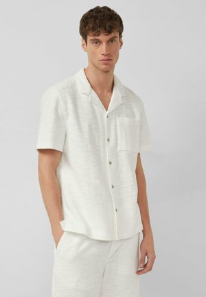 Jeune homme debout les mains dans les poches, portant une chemise blanche texturée à manches courtes boutonnée et un short assorti sur un fond uni.