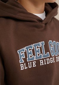 Ruskea huppari, jossa on suuri brodeerattu teksti "FEEL GOOD" sinivalkoisena, varustettu nyöreillä ja pehmeällä, teksturoitulla kankaalla.