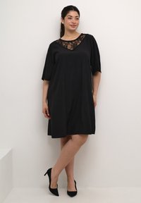 Vestido negro con mangas cortas y sueltas, con un escote de encaje. La tela tiene una textura sutil, combinado con unos tacones altos negros.