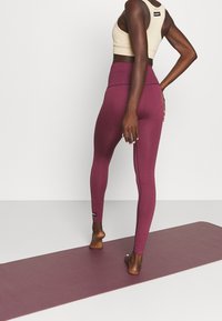 Leggings bordeaux a vita alta con una texture liscia e cuciture laterali, abbinati a un reggiseno sportivo beige realizzato in tessuto elasticizzato.