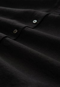 Tissu noir à texture lisse avec des boutons le long de la couture, créant des plis subtils et de la profondeur dans le matériau.