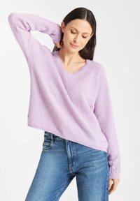 Vrouw in een lichtpaarse V-hals trui met kabelgebreide manchetten en blauwe jeans, poserend met één hand achter haar nek.